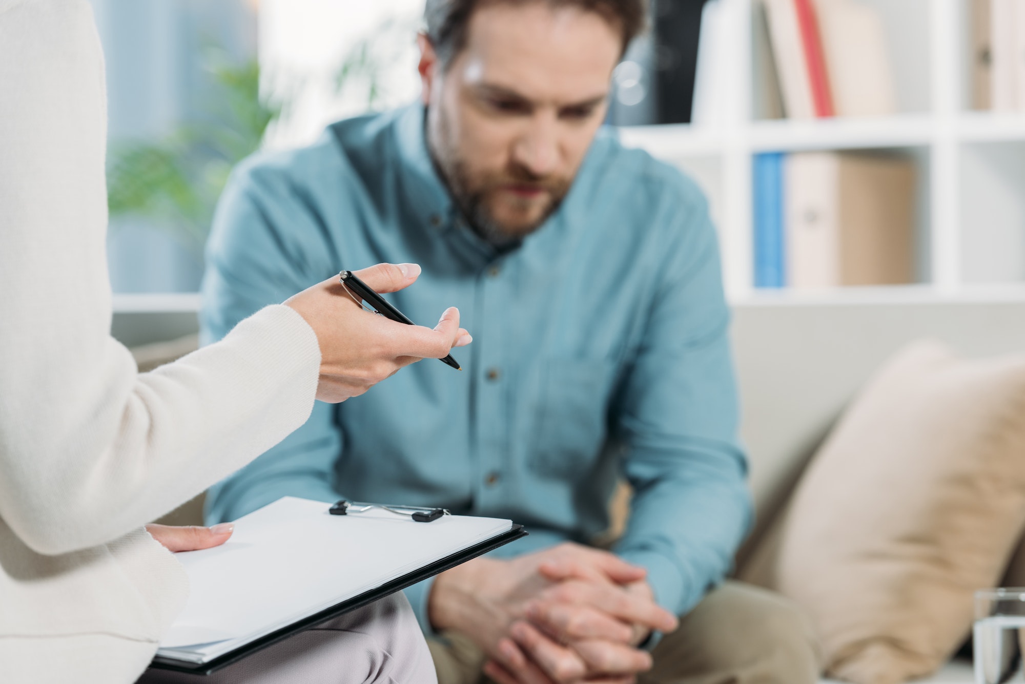 cropped shot of psychotherapist holding pen and clipboard while upset bearded man sitting on couch